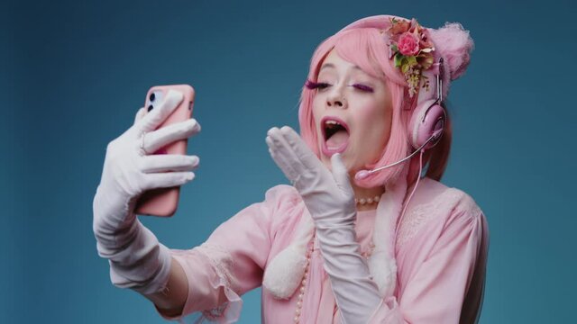 Glamour young sexy candy girl blogger with pink makeup hair and sends an air kiss, takes selfie photo on smartphone on blue background. Internet person grimacing, flirting with subscribers audience.