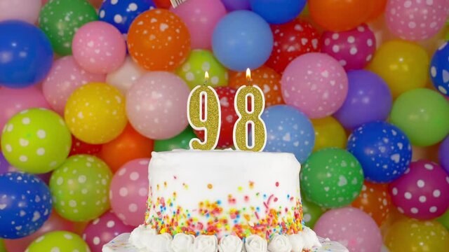 Cake with burning candle number 98. A tasty Birthday Cake on a bright festive background of colorful balloons.