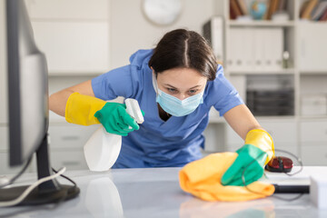 Young woman in a protective mask, cleaning the office with a napkin and detergents, wipes the desktop
