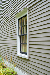 The exterior of a tan color wall of a building. The surface is wood verticle clapboard cape cod siding. There's a vintage multi-pane double hung window in the center with black and white wood trim. 