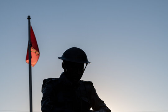 A black silhouette of a soldier standing wearing a military uniform and vintage helmet from the chest up. There's a red flag in the background. The sun is shining through the flag.