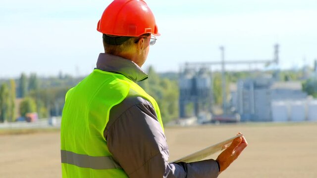 Grain elevator engineer food industry. Mature man engineer with tablet computer checks new agricultural complex with large silo tanks in field