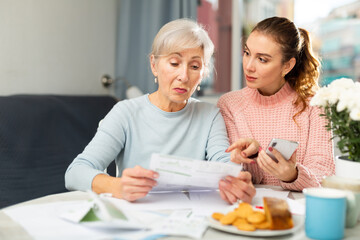 Young woman with smartphone helping grandmother to pay her bills with online payment.