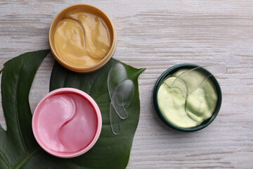 Packages of different under eye patches and tropical leaf on wooden table, flat lay. Cosmetic product