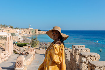 Happy African American woman in yellow dress and sun hat enjoys view of coast of Red Sea on natural background. Panoramic views of blue sea with yachts and coastline, Sharm El Sheikh, Egypt. 