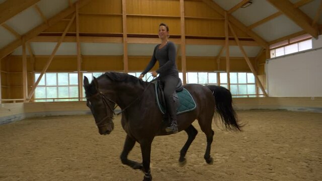 SLOW MOTION: Dark horse prances during flatwork training with happy woman. Energetic horse jumps around the arena during dressage practice with cheerful female rider. Girl rides an energetic mare.