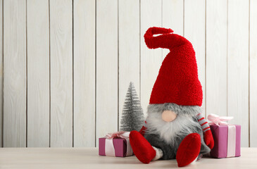 Obraz premium Cute Christmas gnome, gift boxes and small decorative fir tree on table against white wooden background. Space for text