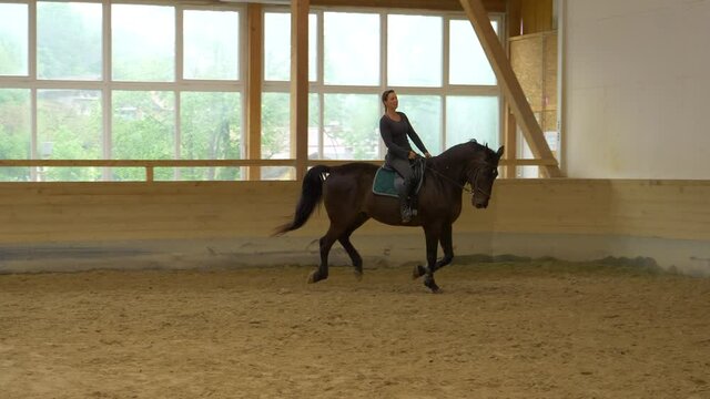 SLOW MOTION: Young Caucasian woman trains her beautiful black horse to gallop. While practicing at an indoor manege. Female horseback rider gallops on her stallion during indoor dressage practice.