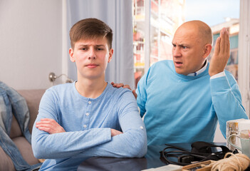 Unhappy teenager boy sitting in home interior while father berating him