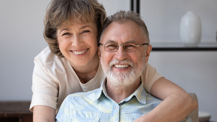 Fototapeta premium Happy senior 50s 60s husband and wife looking at camera with toothy smiles, hugging, laughing. Middle aged mature married couple home head shot banner portrait. Elderly age, relationship concept