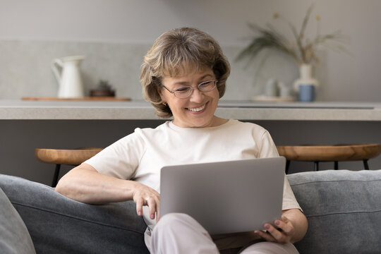 Happy mature 60s woman consulting doctor online, using online telehealth app on laptop, talking on video call, smiling. Older lady chatting on internet, watching funny movie, relaxing on sofa