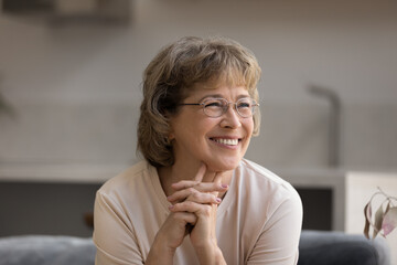 Happy elder 60s woman in glasses sitting on couch at home, looking away with good thoughts thinking...