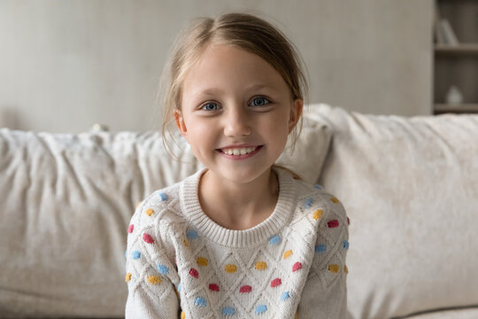 Head shot portrait of smiling adorable little kid girl sitting on comfortable couch, holding distance video call conversation, web camera view. Joyful cute small child posing alone in living room.