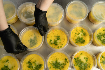 many disposable plates with soup in a lunch box stand on the table in the kitchen in a restaurant female hands are covering the food with a lid. Takeaway food concept during quarantine or pandemic.