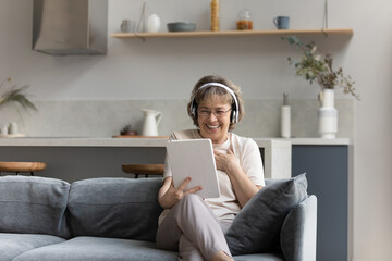 Cheerful elderly 60s woman in wireless headphones watching movie, media content, webinar on tablet...