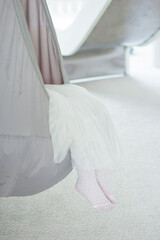 Close up girls legs in white tights and white dress. Cropped view of little girl relaxing in hanging grey chair bag or hammock in light room with carpet. Swing at home.