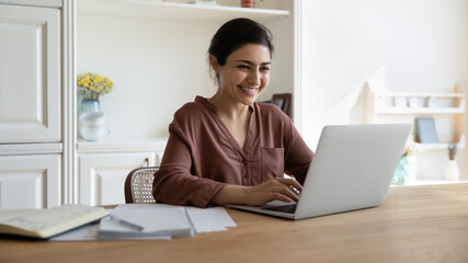 Happy millennial indian businesswoman working on computer in modern home office, checking email,...