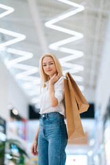 Vertical portrait of attractive blonde young woman holding shopping paper bags with purchases standing in hall of mall centre, blurred background. Portrait of pretty shopaholic lady posing at store.