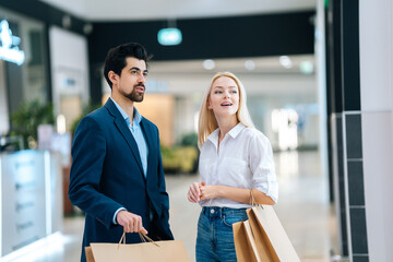 Beautiful happy young couple holding shopping paper bags with purchases and looking upon showcase at mall. Handsome bearded man and attractive blonde woman purchasing together at store centre.