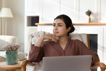Thoughtful beautiful young indian ethnic woman considering problem solution, planning weekend or vacation, working in computer remotely, sitting in comfortable armchair in modern living room.