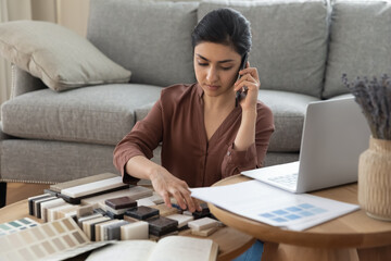 Concentrated young indian woman sitting at table with swatches, discussing interior design with professional designer by phone, making order choosing materials for decorations at home, planning repair