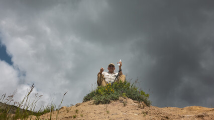 A man is sitting on top of a hill, against a cloudy sky. In his hands is an open bottle of water, a hiking stick. Around lush green vegetation, wildflowers. Kamchatka