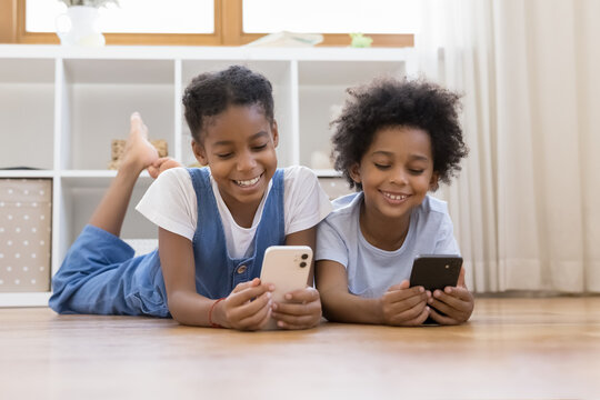 Sweet African sibling children relaxing on warm floor, enjoying leisure time with digital gadgets, using learning application on mobile phones, chatting on social media, watching online media content