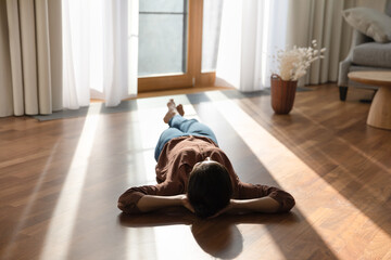 Relaxed happy young indian ethnicity woman homeowner lying alone on warm wooden floor with underfloor heating, enjoying carefree peaceful weekend leisure time alone in modern stylish living room. - obrazy, fototapety, plakaty