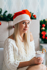 Blonde woman wearing white knitted dress Santa hat sit on bed in Christmas-decorated bedroom and drink hot cocoa with marshmallows.