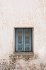 Fototapeta na wymiar White wall with blue old wooden window with closed shutters, Stone wall with rough and cracked surface, vertical format 