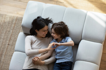Overhead view loving Asian mother and little son lying on sofa join their fingers showing heart...