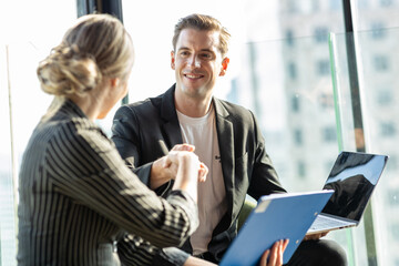 Young business man and young business woman taking to each other They are in white shirt and suit. They are in a hotel lobby. The view behind is cityscape.