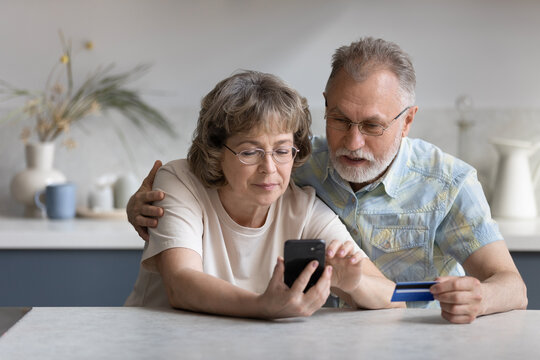 Serious older married couple shopping on internet stores, using banking, ecommerce app on cellphone, paying for purchase by credit card, making transaction, transfer, money order. Finance concept