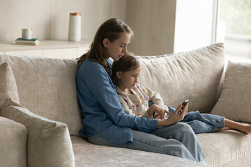 Happy beautiful millennial mother embracing sincere cute kid daughter, learning using software cellphone applications, web surfing information, shopping in internet store, resting on cozy sofa.