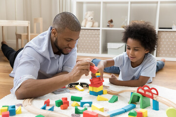 Loving young African dad and cute little son engaged in game with construction cubes, railway, resting and playing on warm floor, talking, enjoying playtime activities together. Family, fatherhood