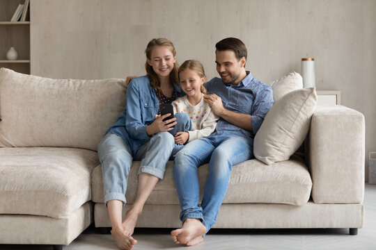 Full length bonding Caucasian two generations family using smartphone, resting on cozy sofa in modern living room. Addicted to modern tech happy young couple parents and little daughter playing games.
