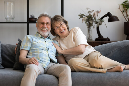 Happy older 60s couple relaxing in living room home portrait. Senior husband and wife resting, hugging on couch, looking at camera, laughing, smiling, talking on video call. Screen view