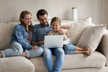 Happy young Caucasian couple parents using computer applications with laughing small cute kid daughter, enjoying playing online games, choosing goods shopping in internet store, watching cartoons.