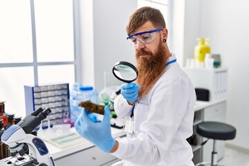 Young redhead man wearing scientist uniform holding marihuana weed with loupe at laboratory