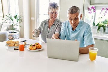 Senior caucasian couple having breakfast using laptop at the kitchen.