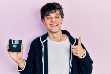 Handsome hipster young man holding floppy disk smiling happy and positive, thumb up doing excellent...