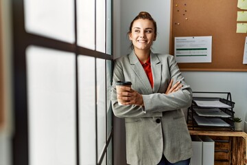 Young woman business worker drinking coffee standing with arms crossed gesture at office