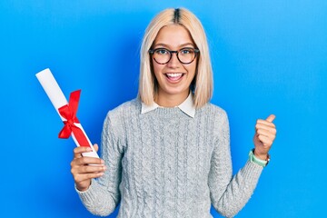 Beautiful blonde woman holding graduate degree diploma pointing thumb up to the side smiling happy with open mouth