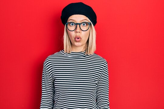 Beautiful blonde woman wearing french look with beret an glasses scared and amazed with open mouth for surprise, disbelief face