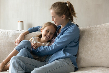 Caring millennial mother tickling adorable small kid daughter, having fun together resting on couch, enjoying spending weekend leisure time together, showing sincere multigenerational family relations