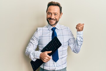 Middle age businessman holding keyboard pointing thumb up to the side smiling happy with open mouth