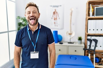 Middle age physiotherapist man working at pain recovery clinic sticking tongue out happy with funny expression. emotion concept.