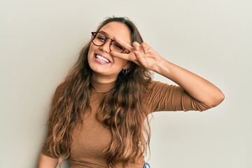 Young hispanic girl wearing casual clothes and glasses doing peace symbol with fingers over face, smiling cheerful showing victory