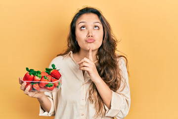 Young hispanic girl holding strawberries thinking concentrated about doubt with finger on chin and...