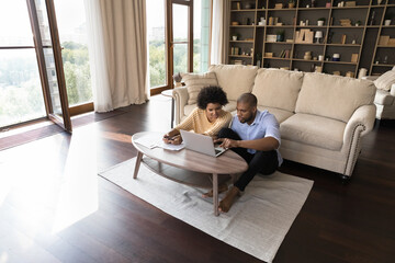 Young African American married couple maintaining family budget, using finance app on laptop computer, calculating money, paying paper bills, invoice, doing domestic paperwork at home. Wide shot